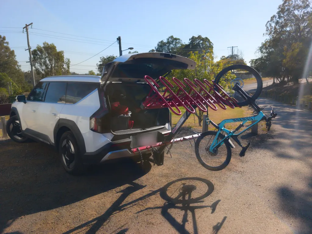A white Kia EV9 parked on a gravel verge with the tailgate up and a red vertical bike rack on the tow bar holding one turquoise mountain bike.