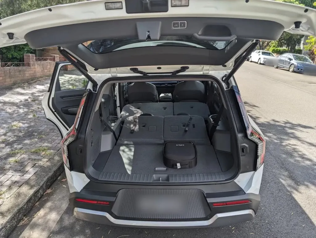 The back of a white Kia EV9 with the tailgate up and both rear seat rows folded flat; the number plates are blurred.