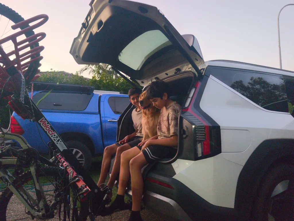 Two boys and a golden retriever sitting in the open boot of a white Kia EV9 in a driveway at dusk, with mountain bikes on a rack beside the car.
