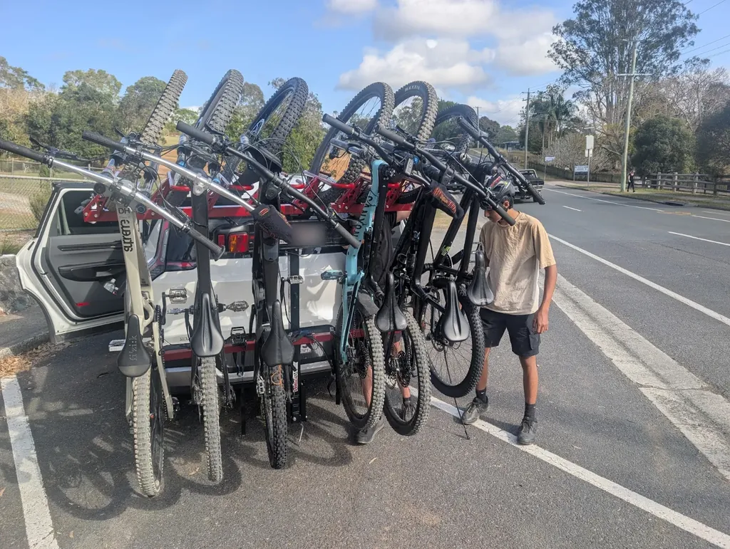 Six mountain bikes hanging wheels-on from a vertical rack on the back of a white EV9 at the roadside, with a young rider in a helmet checking one of them.